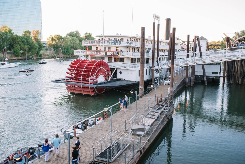 A classic paddlewheel riverboat, "Delta King Hotel," docked on a serene river with people on the pier. Trees and a glass building visible in the background.