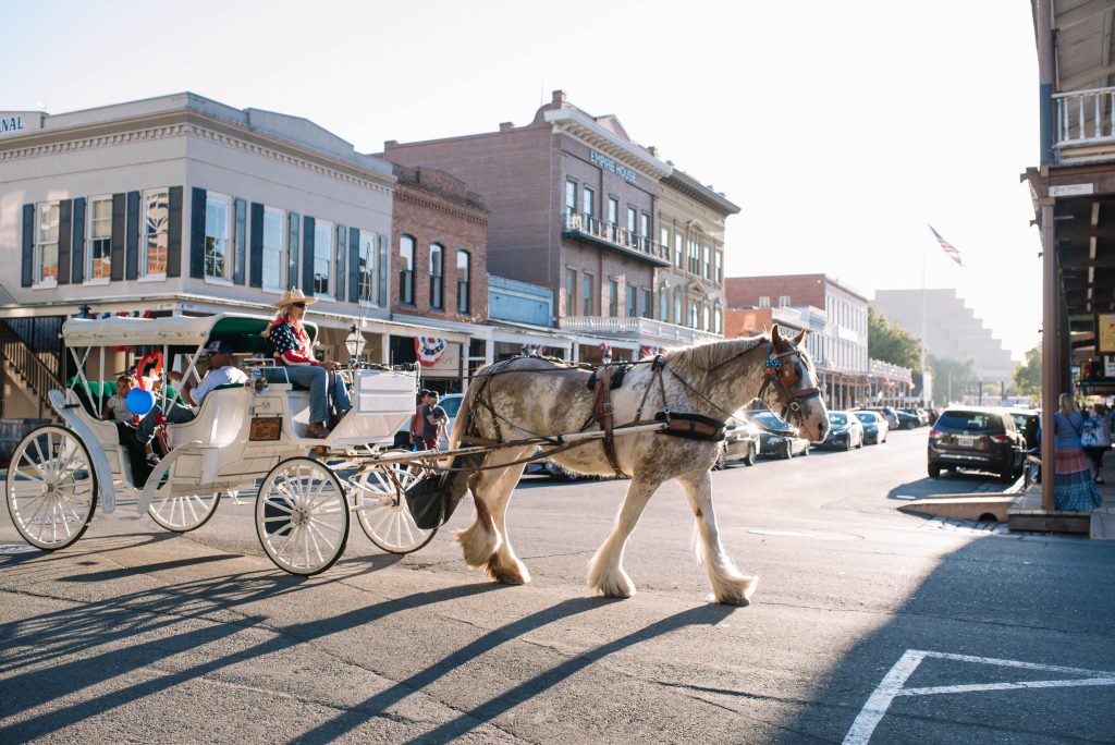 A horse-drawn carriage with two passengers rides through a sunny, historic town street. The scene feels nostalgic with its old brick buildings.