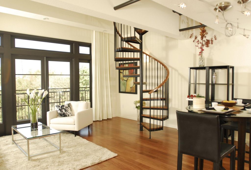 Modern living room with a spiral staircase. Includes a white armchair, glass coffee table with lilies, and dining area, on warm wood flooring. Bright and airy ambiance.