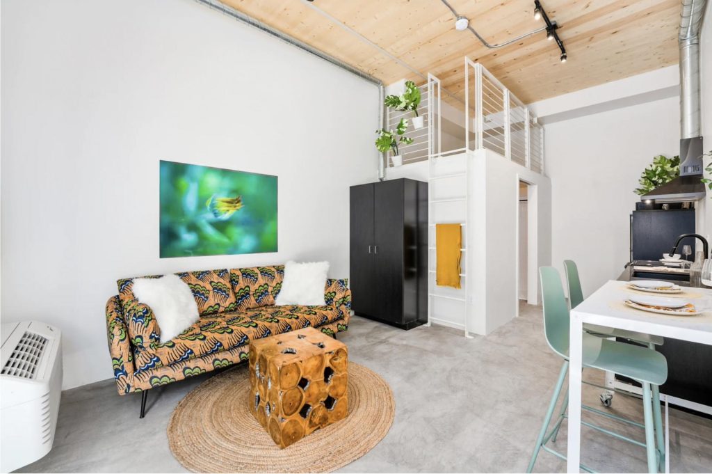 Stylish modern living room with a colorful patterned sofa and white cushions. A unique wood block coffee table sits on a woven rug. A vibrant nature photo hangs on a white wall. There's a black wardrobe, loft with plants, and kitchenette with stools, conveying a cozy, contemporary feel.