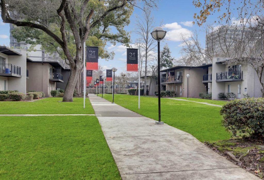 Pathway through a tranquil apartment complex, bordered by lush green lawns and mature trees. Banner flags line the path, with modern buildings on each side under a blue sky.