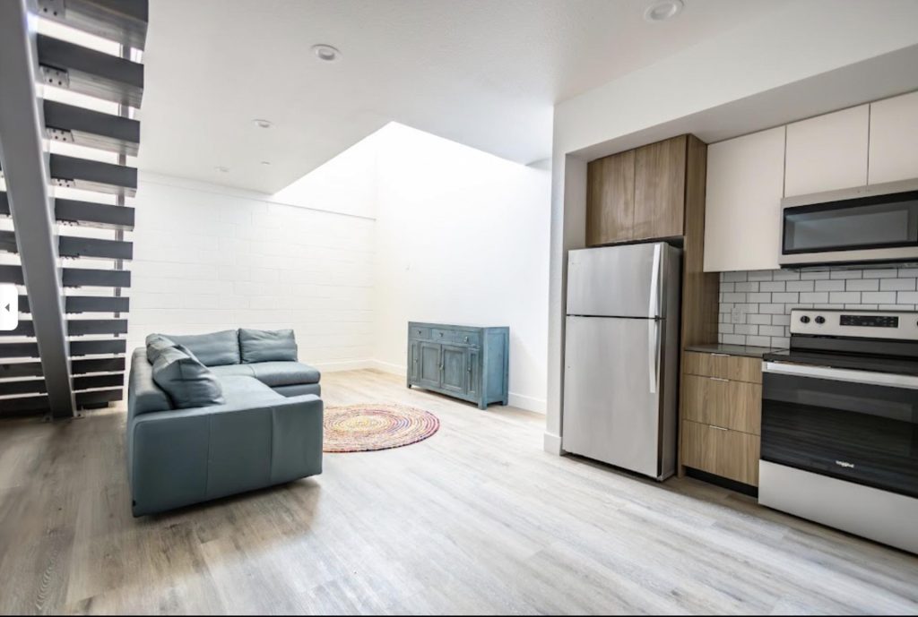 Modern loft interior with a gray couch, blue sideboard, and a colorful round rug. Bright lighting, open kitchen with stainless steel appliances. Minimalist and airy.