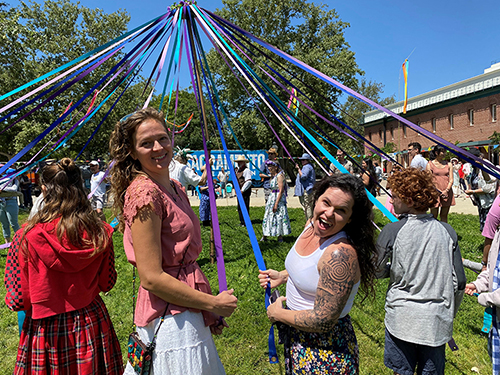 A festive outdoor gathering features people holding colorful ribbons tied to a maypole, smiling and enjoying the sunny day, surrounded by trees and buildings.