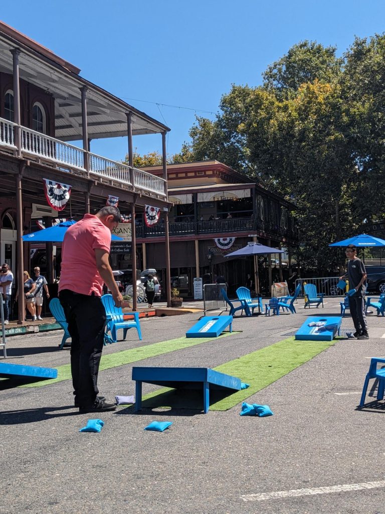 A man in a pink shirt plays cornhole on a sunny day, set in a street lined with historic buildings and decorated with banners. Blue chairs and umbrellas add to the relaxed, festive atmosphere.