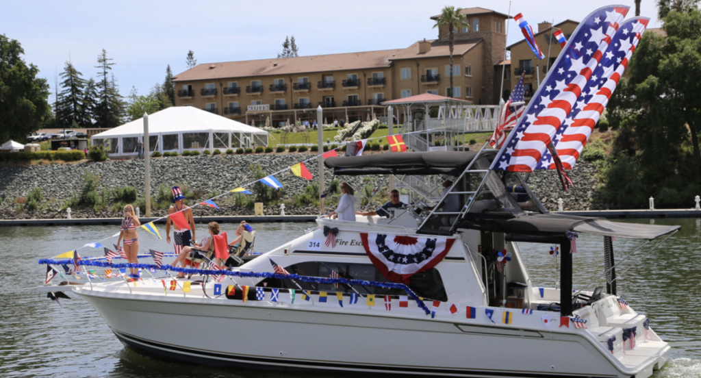 Boat decorated with colorful flags and large American flag sails, cruising on a lake. People in festive attire wave, creating a joyful, celebratory atmosphere.
