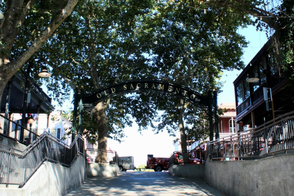 A sunny day in Old Sacramento, featuring a large archway with the name above a tree-lined path. Vintage buildings line the sides, creating a historic ambiance.