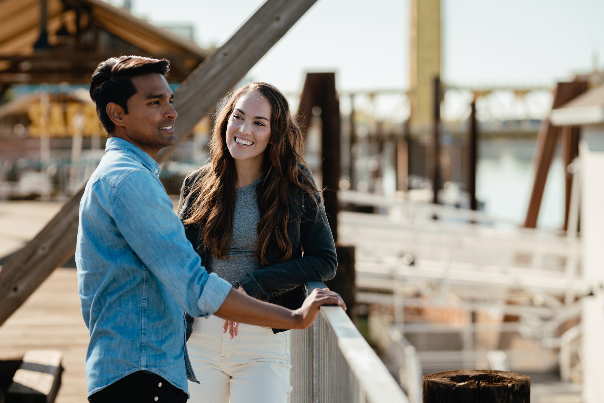 A man in a denim shirt and a woman in a cardigan smile at each other on a sunny waterfront boardwalk, conveying warmth and friendly conversation.