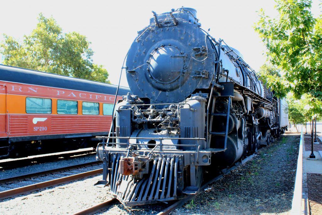 A black vintage steam locomotive is displayed on train tracks beside an orange passenger car labeled "Southern Pacific." Sunlight filters through nearby trees.