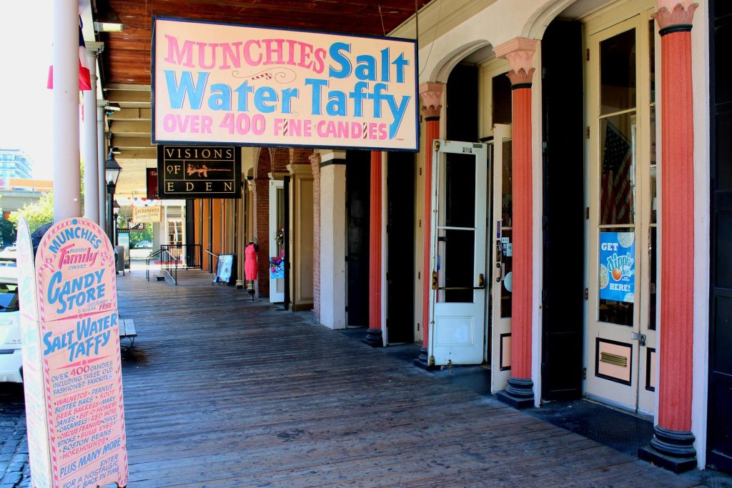 Cozy candy store entrance with a pink and blue "Munchies Salt Water Taffy" sign. Rustic wooden porch, inviting open doors, vibrant, nostalgic feel.