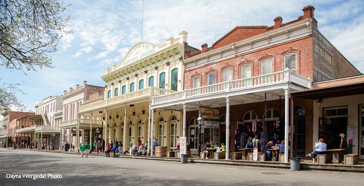 A lively historic street with colorful vintage buildings features people walking and sitting. The blue sky and vibrant architecture evoke a cheerful atmosphere.