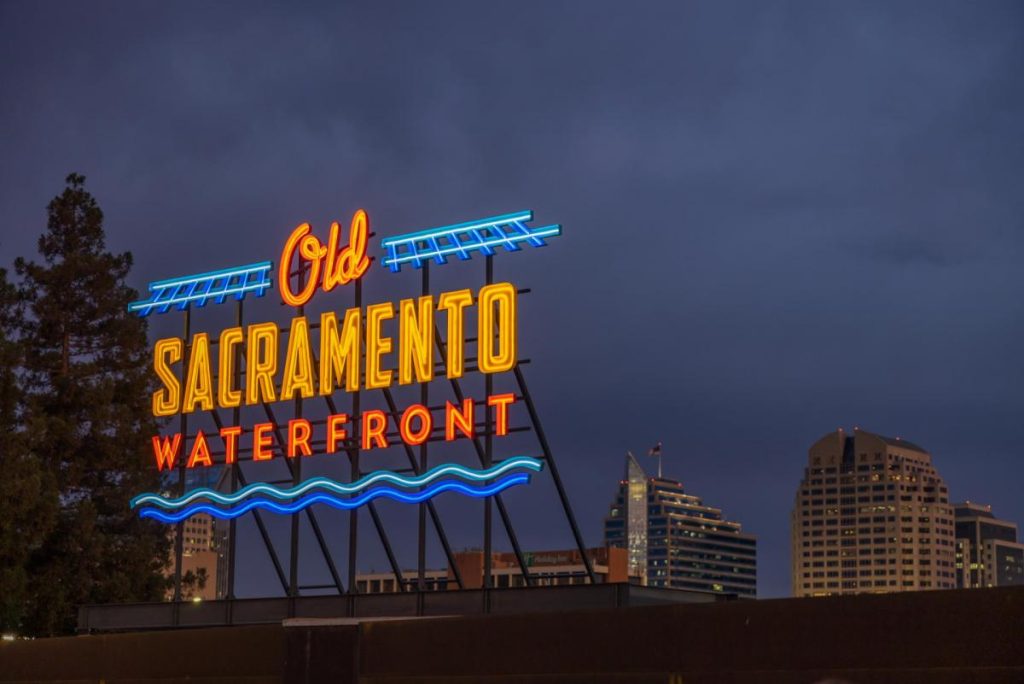Neon sign reading "Old Sacramento Waterfront" glows in red, yellow, and blue against a cloudy night sky, with a city skyline in the background.