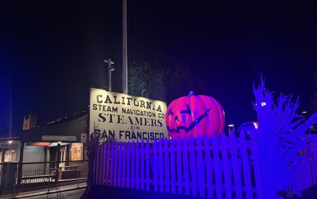 A glowing jack-o'-lantern rests beside a sign reading "California Steam Navigation Co. Steamers for San Francisco," surrounded by a white picket fence under blue lighting, creating an eerie, festive Halloween atmosphere.