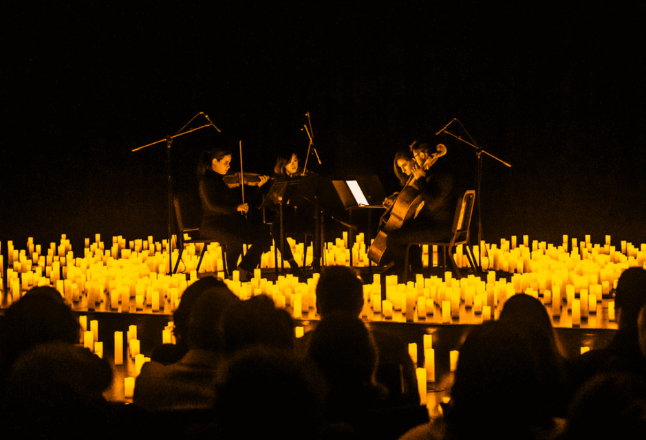 A string quartet performs surrounded by glowing candles in a dark room. The warm yellow light creates an intimate, serene atmosphere for the audience.