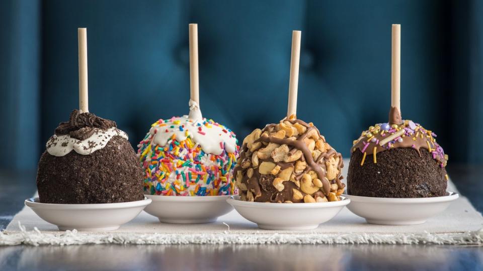 Four gourmet candy apples in small dishes are arranged in a row. Each is coated in toppings: crushed cookies, rainbow sprinkles, nuts, and chocolate drizzle. A cheerful and indulgent display.