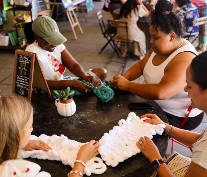 Four women sit around a table knitting, their expressions focused and content. A small plant and drink menu are on the table, creating a cozy, communal atmosphere.