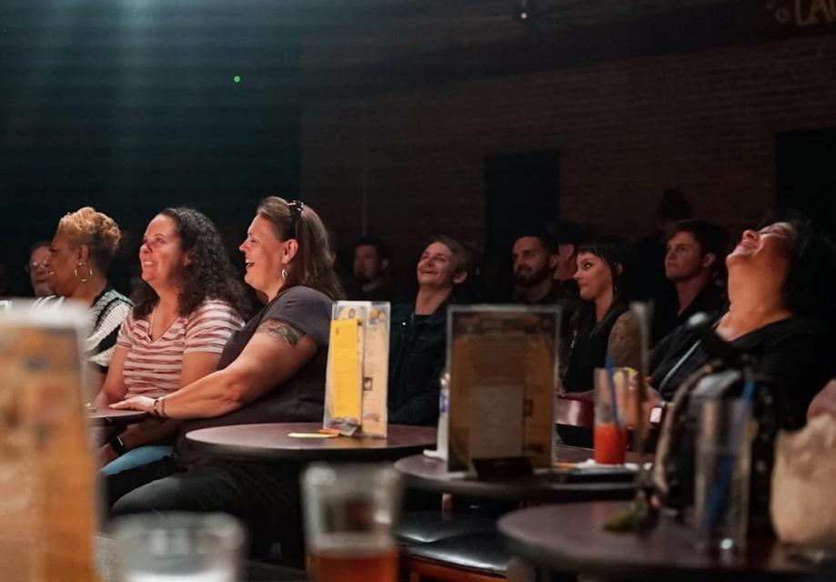 Audience members at Laughs Unlimited laugh and smile warmly during a live comedy show in a dimly lit venue. Tables hold drinks and menus, highlighting a relaxed atmosphere.